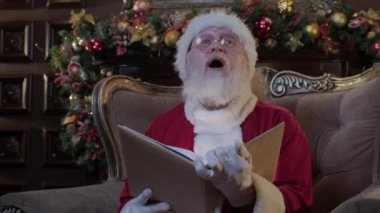 Yeni yıl kostümlü, gri sakallı Noel Baba koltukta oturur ve peri masallarıyla sihirli kitaplar okur. Noel Baba 'nın yüzüne yansıyan ışık huzmeleri arka plandaki Noel ağacına vurur.