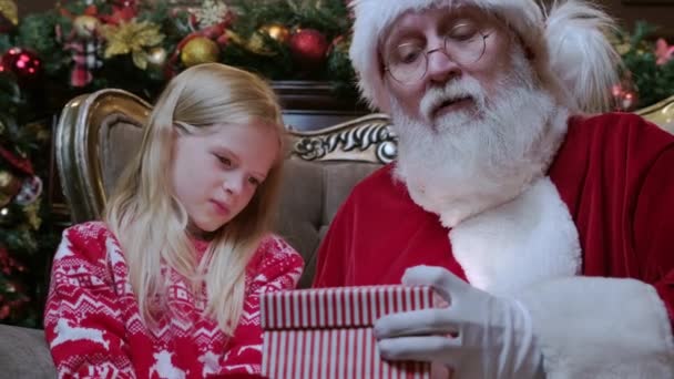 Santa Claus dává dárek magie box dívka, jasné světlo svítí z krabice na malou blondýnu sedí v křesle v pokoji s krbem a vánoční stromeček zdobené v pokoji. — Stock video