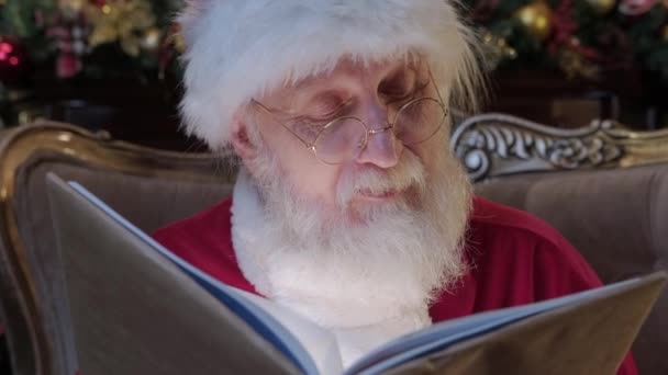 Zbliżenie Świętego Mikołaja w okularach z szarą brodą w nowym roku kostium siedzi na krześle i czytania magicznej książki z bajki Boże Narodzenie, promienie światła Blask od bogini na twarzy Świętego Mikołaja. — Wideo stockowe