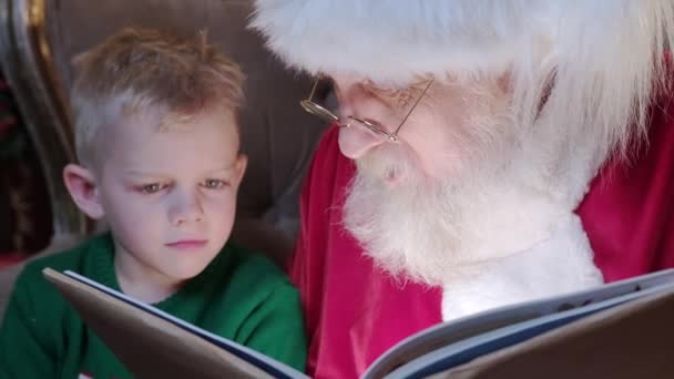 Snäll gubbe med långt grått skägg Jultomten läser julhistoria till liten blond pojke sitter i stol i rum med öppen spis och julgran dekorerad. Begreppet nyår semester. — Stockvideo