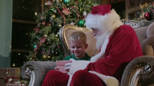 Santa Claus dává kouzelné dárkové krabice chlapec, jasné světlo svítí z krabice na malé blonďaté dítě sedí v křesle v pokoji s krbem a vánoční stromeček zdobené v pokoji. — Stock video
