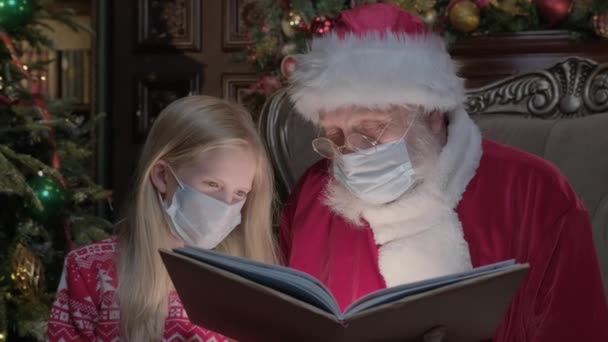 Santa Claus v lékařské masce hostující dívku během epidemie koronaviru, Santa čte magickou knihu s jasným světlem svítí na ně. Dívka v období coronavirus čtení knihy s Santa. — Stock video
