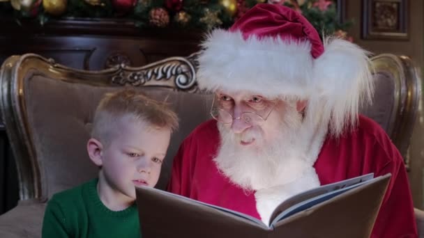 Vrai père Noël vieil homme avec une longue barbe grise, lit l'histoire de Noël au petit garçon assis dans la chaise dans la chambre. Boy lit le livre avec Saint Nicolas à Noël. Bonne année et jour de Noël — Video