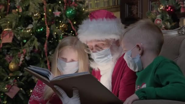 Światowa pandemia koronawiry19, dzieci brat i siostra czytają magiczną noworoczną opowieść z Mikołajem w masce medycznej, siedzącym obok niego na krześle w pokoju z wnętrzem noworocznym. — Wideo stockowe