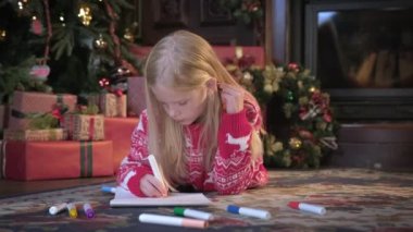 Kırmızı kazaklı sarışın kız Noel ağacının yanında yerde yatıyor ve Noel Baba 'ya keçeli kalemlerle mektup yazıyor. Çocuk, Noel Baba için dileklerle mektup yazıyor.