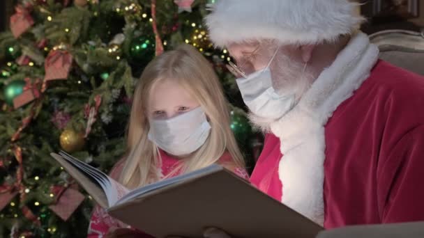 Originální Santa Claus spolu s malou dívkou v koronavirové virové pandemii, sedět v lékařské masce spolu a číst pohádky v magické knize s jasnou září světla. Vánoční kouzlo Vánoc — Stock video