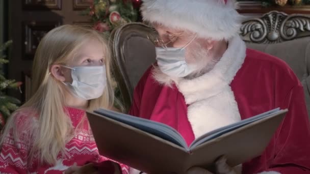 Äkta jultomten i skyddsmask från virus läser magisk bok med blond flicka. Ljus från boken lyser i ansiktet på flicka och jultomten läser julberättelser i mörkt rum — Stockvideo