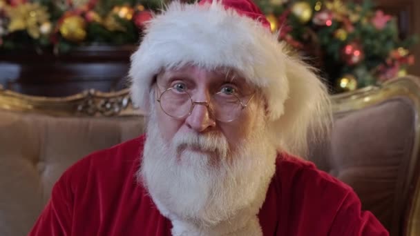 Portrait of a good authentic Santa Claus wearing a hat and a new years costume, grandfather looks at the camera and adjusts his gold glasses. the concept of the Christmas spirit closeup — Stock Video