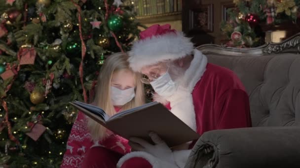 Santa Claus v lékařské masce hostující dívku během epidemie koronaviru, Santa čte magickou knihu s jasným světlem svítí na ně. Dívka v období coronavirus čtení knihy s Santa. — Stock video
