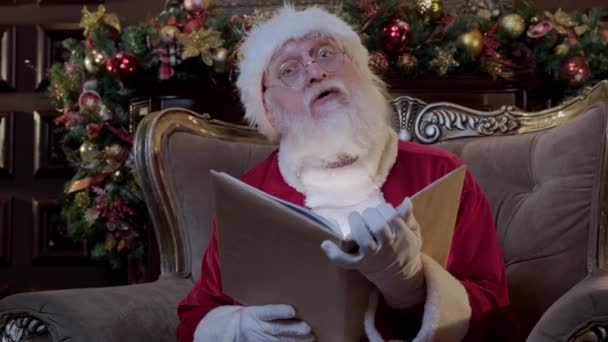 聖ニコラスは彼の家のアームチェアに座っている間に輝く開いた魔法の本を読んでいます。サンタクロースは箱の中の子供たちにおとぎ話を読み、サンタの祖父から新年の挨拶. — ストック動画