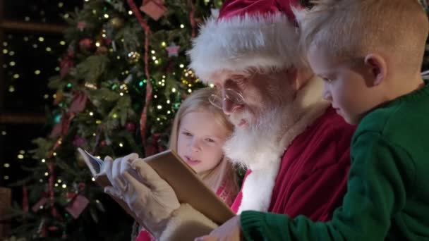 Dzieci brat i siostra czytali noworoczną opowieść z Mikołajem siedzącym obok niego na krześle w pokoju z wnętrzem noworocznym. Święty Mikołaj czyta bajki Boże Narodzenie do małych dzieci, chłopiec i dziewczyna — Wideo stockowe