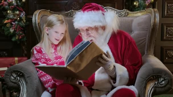 Pravý Santa Claus otevírá a čte kouzelnou knihu na Štědrý večer s blondýnou sedí v náručí svého dědečka. Světlo z knihy svítí na tvář dívka a Santa čtení vánoční příběhy v pokoji — Stock video