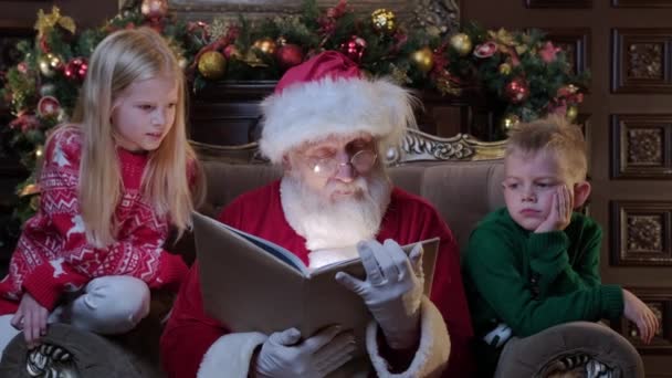 Дети брат и сестра читают новогоднюю сказку, а Санта Клаус сидит рядом с ним в кресле в комнате с новогодним интерьером. Санта-Клаус читает рождественскую сказку маленьким детям, мальчику и девочке. — стоковое видео