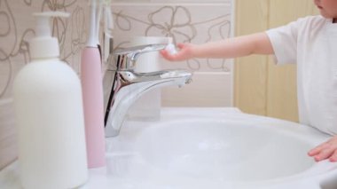 Çocuk elinde sabun ve köpük tutuyor musluğa getiriyor ve suyun altında ellerini yıkıyor, banyoda diş fırçası ve şişelerin arka planına karşı suyu kapatıyor.
