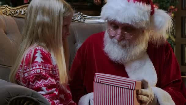 Santa Claus gives gift magic box girl, bright light shines from box to little blonde girl sitting in chair in room with fireplace and Christmas tree decorated in room. — Stock Video