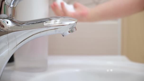 Primer plano niño recoge el jabón y la espuma en sus manos lo lleva al grifo y bajo el chorro de agua se lava las manos, cierra el agua en el baño contra el fondo de cepillos de dientes y botellas. — Vídeos de Stock