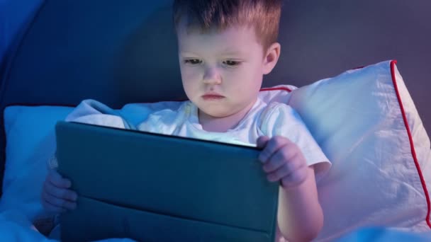 Menino loiro pequeno à noite na cama assistindo vídeo no tablet dispositivo móvel. Kid assiste desenhos animados antes de ir para a cama, sentado na cama em raios de lua e lâmpada. — Vídeo de Stock