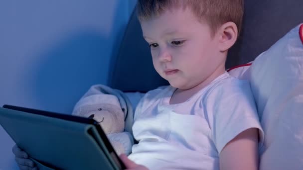 Pojken ansikte upplyst från bärbar skärm medan du spelar eller lära sig via globalt Internet. Porträtt av söt blond kaukasier pojke tittar på tecknad tablett sitter i sängen i månsken från fönstret hemma. — Stockvideo