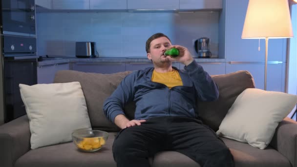 El hombre gordo sin afeitar come papas fritas y bebe cerveza alcohólica, se sienta relajado en el sofá en casa y ve la televisión, cambia de canal con control remoto, ríe y disfruta de su descanso. — Vídeos de Stock