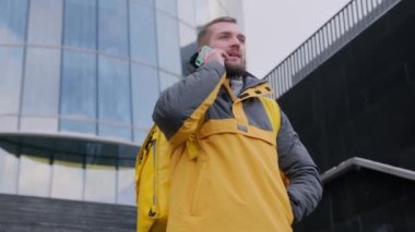 Kurye caddede teslimat adresi arıyor. Akıllı telefonu olan yakışıklı bir adam açık havada sarı termosta yemek dağıtıyor. Restoran yemeği getiren adamın portresi. Sipariş getiriyor..