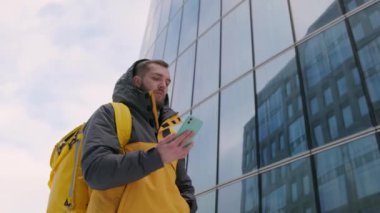 Sırtında sarı bir sırt çantası olan traşsız kurye kışın caddede uzun cam binaların arasında yürüyor, telefonla şehri dolaşıyor ve teslimat adresini buluyor. Kurye teslimatı kavramı.