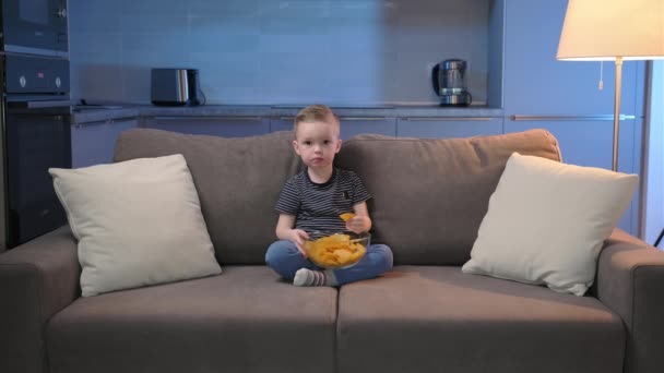 El niño se sienta en la cama en la habitación y enciende la televisión, come papas fritas. El chico cambia de canal en la TV. Ver dibujos animados o películas de niños. Lindo niño en casa sin padres viendo la televisión. — Vídeo de stock