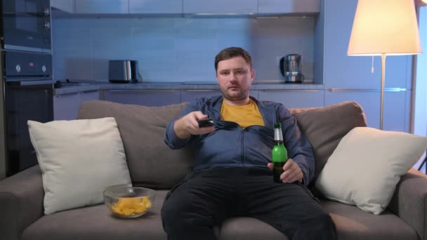 Urychlené video Muž sleduje zábavné televizní pořady na televizní obrazovce, pije alkohol a jíst. Neoholený muž sedí v noci na pohovce v tmavém obývacím pokoji a dívá se na televizi, jí brambůrky a pije pivo. — Stock video