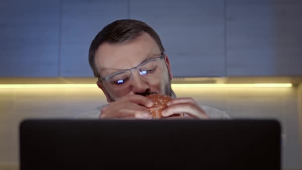 Retrato de hombre hambriento, sin afeitar con gafas comiendo hamburguesa grande con carne de res de restaurante de comida rápida y viendo la película en la pantalla del ordenador portátil en la habitación oscura contra el fondo de la cocina. — Vídeos de Stock