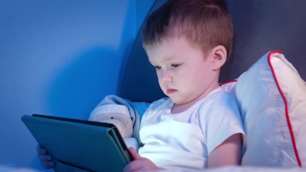 Akşamları yakışıklı, beyaz tenli sarışın çocuk, karanlık çocuk odasında oyuncak ayıyla birlikte tablette çizgi film izliyor. Childs 'ın yüzü dizüstü bilgisayardan gelen ışıkla aydınlatılır, evde boş zaman geçirilir.. — Stok video