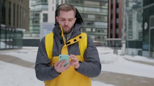 Entrega homem com mochila de costas em uniforme andando rua abaixo no inverno entre edifícios altos, se comunicando com o cliente no telefone, navegando através do telefone. Conceito de entrega de correio. — Vídeo de Stock