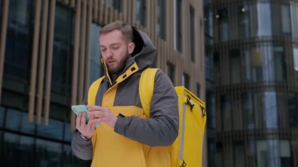 Sırtında sarı termo çantalı, traşsız beyaz kurye teslimat adresini arıyor. Müşteri teslimatı beklerken telefonla konuşuyor. Şehirde fast food servisi.. — Stok video