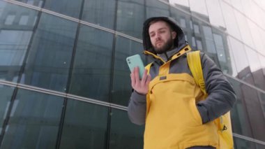 Sırtında sarı bir sırt çantası olan traşsız kurye kışın caddede uzun cam binaların arasında yürüyor, telefonla şehri dolaşıyor ve teslimat adresini buluyor. Yavaş çekim videosu.