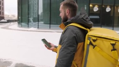 Fast food restoranından hızlı teslimat. Teslimatçı, yüksek binalar arasında caddede adres aramak için cep telefonu kullanıyor. Kurye soğuk havada termos çanta ve akıllı telefonla sokakta yürür..