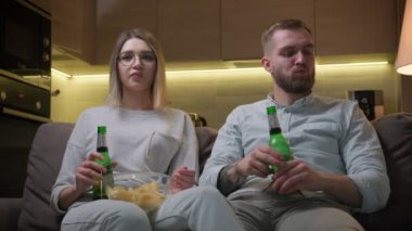 Tıraşsız bir adam ve gözlüklü bir kız evdeki koltukta oturup alkollü bira içip patates cipsi yiyorlar. TV 'de kız ve erkek dizisi ya da film izlemek..