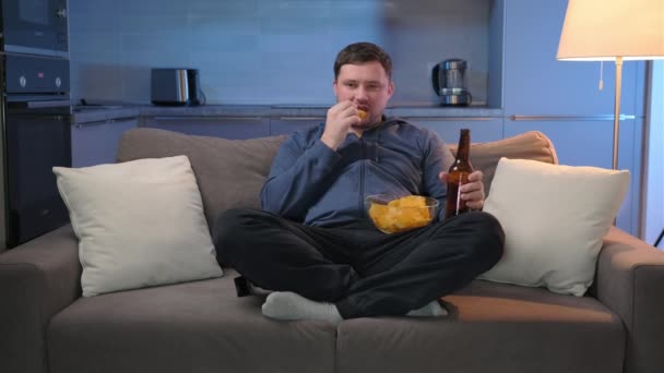Homem sem barba senta-se à noite em um sofá em uma sala de estar escura, assistindo TV, comer batatas fritas e beber cerveja, rindo alegremente assistindo a um programa de entretenimento programa de TV. Lazer noturno em casa. — Vídeo de Stock