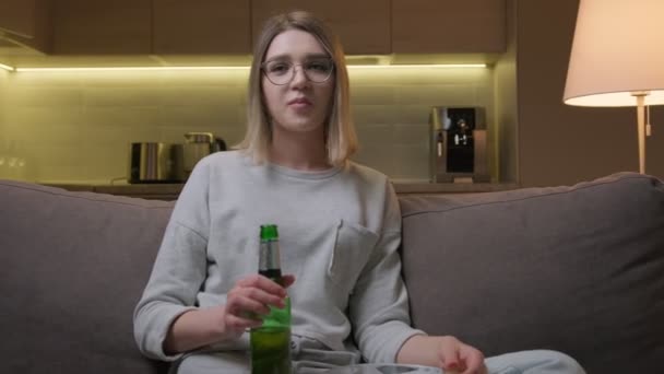 Chica rubia caucásica con gafas ve noticias o programas de televisión o película en la televisión bebe cerveza y come patatas fritas sentadas en el sofá en casa por la noche. Mujer ve programas de televisión o competiciones deportivas en la televisión. — Vídeos de Stock