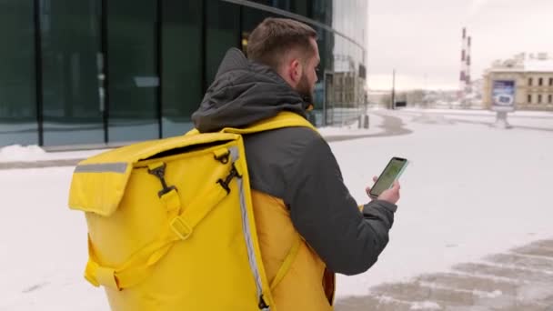 Futár sétál termosz táska és okostelefon az utcán hideg időben. A futár mobiltelefont használ, hogy az utcán keressen címet magas épületek között. Gyors szállítás gyorskajáldából. — Stock videók