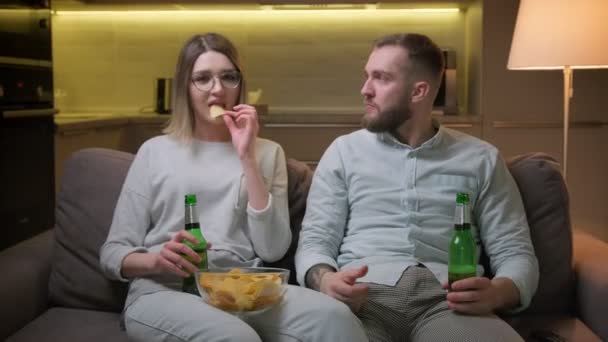 Jovem bonito caucasiano cara e menina assistir a um filme interessante na TV, juntamente com beber cerveja e comer batatas fritas na sala de estar escura no sofá. — Vídeo de Stock