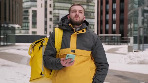 Şehirdeki termos çantalı sarı üniformalı traşsız kurye kışın iş merkezinde restorandan müşteriye yemek dağıtıyor. Ciddi bir adamın portresi restorandan telefonla yemek getiriyor.. — Stok video