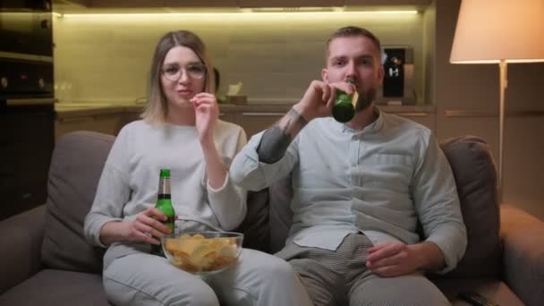 Portrait tourné de jolie jeune femme blonde et un homme non rasé, fans de football regarder match à la télévision dans le salon, boire de la bière alcoolisée et manger des croustilles. — Video