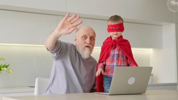 Abuelo anciano con barba gris, junto con el nieto-un niño en impermeable rojo, sentarse en el ordenador, comunicarse a través de un enlace de vídeo con los familiares, mirar a la cámara para el operador y agitar las manos con. — Vídeo de stock