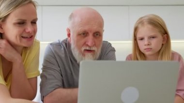 70 yaşında yaşlı, kır saçlı, yaşlı bir emekli modern dizüstü bilgisayar kullanmayı öğreniyor. Gülümseyen genç bir kadın ve kızı yaşlı bir babaya evde dinlenirken laptopta uygulamaları nasıl kullanacağını gösteriyor..