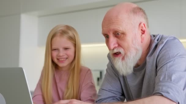 祖父と小さな孫娘使用してノートパソコン一緒に座ってテーブル,二世代の孫娘女の子持っています楽しいです古いです祖父の腕時計漫画ビデオ通話自宅でPC画面を見ます — ストック動画