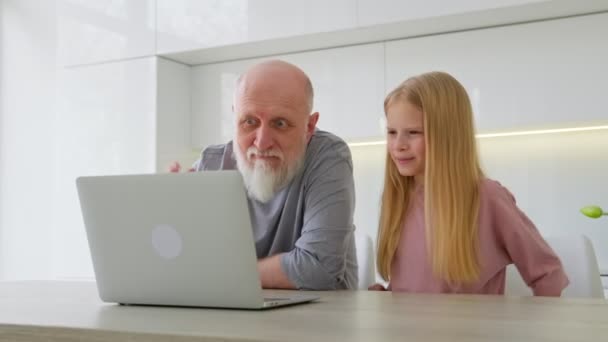 Comunicación familiar a distancia a través de vídeo. Anciano abuelo de pelo gris y su nieta se sientan en el otro lado de la computadora y se comunican a través de enlace de vídeo, riendo y agitando las manos. — Vídeo de stock