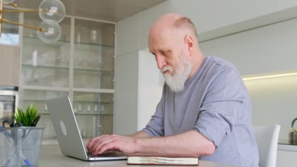 Velho idoso bonito trabalha em casa atrás do laptop. Avô aposentado aprende a usar teclado laptop e posar enquanto está sentado em casa no local de trabalho. Nova tecnologia para idosos. — Vídeo de Stock