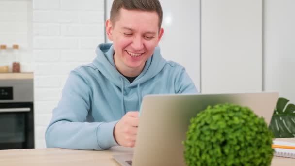 Un uomo lavora a casa da remoto, lavora fuori dall'ufficio. Giovane uomo caucasico lavora a casa da lontano, si siede a casa al computer con computer portatile e scrive messaggi in chat sul telefono. — Video Stock