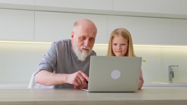 Anciano abuelo, junto con la nieta rubia, pregunta y aprende a usar aplicaciones informáticas. Enfocado chica joven explicando software portátil a un abuelo jubilado interesado. — Vídeo de stock