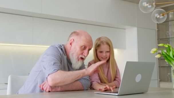 O avô de cabelos grisalhos idoso e sua neta se sentam no outro lado do computador e se comunicam via link de vídeo, rindo e acenando com as mãos. Comunicação familiar à distância através do vídeo. — Vídeo de Stock