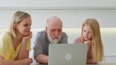 Birkaç nesildir arkadaş canlısı üç aile birlikte dizüstü bilgisayar kullanıyor. Mutlu büyükbaba, genç yetişkin anne ve kız torun evde bilgisayardaki bir programı izlerken gülüyorlar..