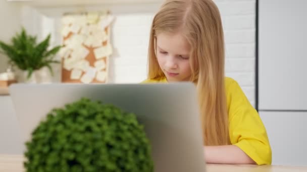 ブロンドの女子高生はノートパソコンを使ってオンラインで学習し、ノートブックにメモを取る。十代の女子高生は自宅のテーブルに座りながらインターネットビデオコースを見ています。距離学習の概念. — ストック動画